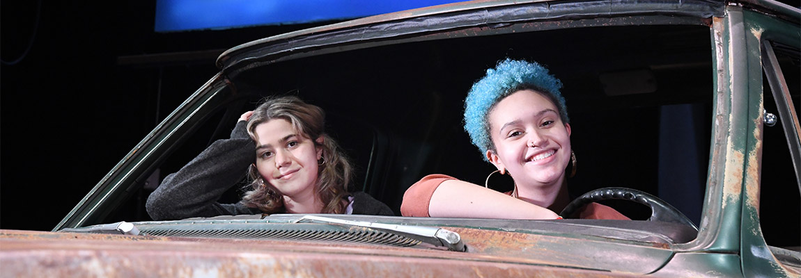 Actors Marina Kunas '25 and Bridget Wiggan '23 look through the winshieled on a battered truck on the set of